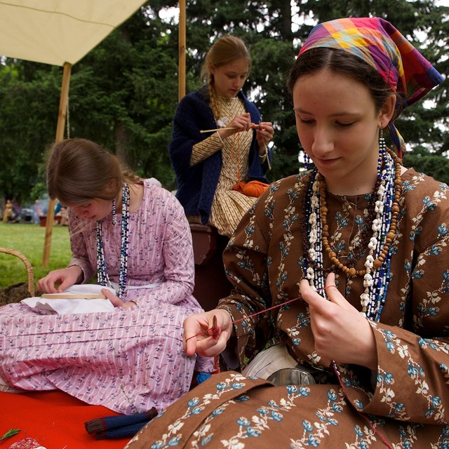 Women dressed in 1840s style clothing do handwork at an event. NPS photo. 