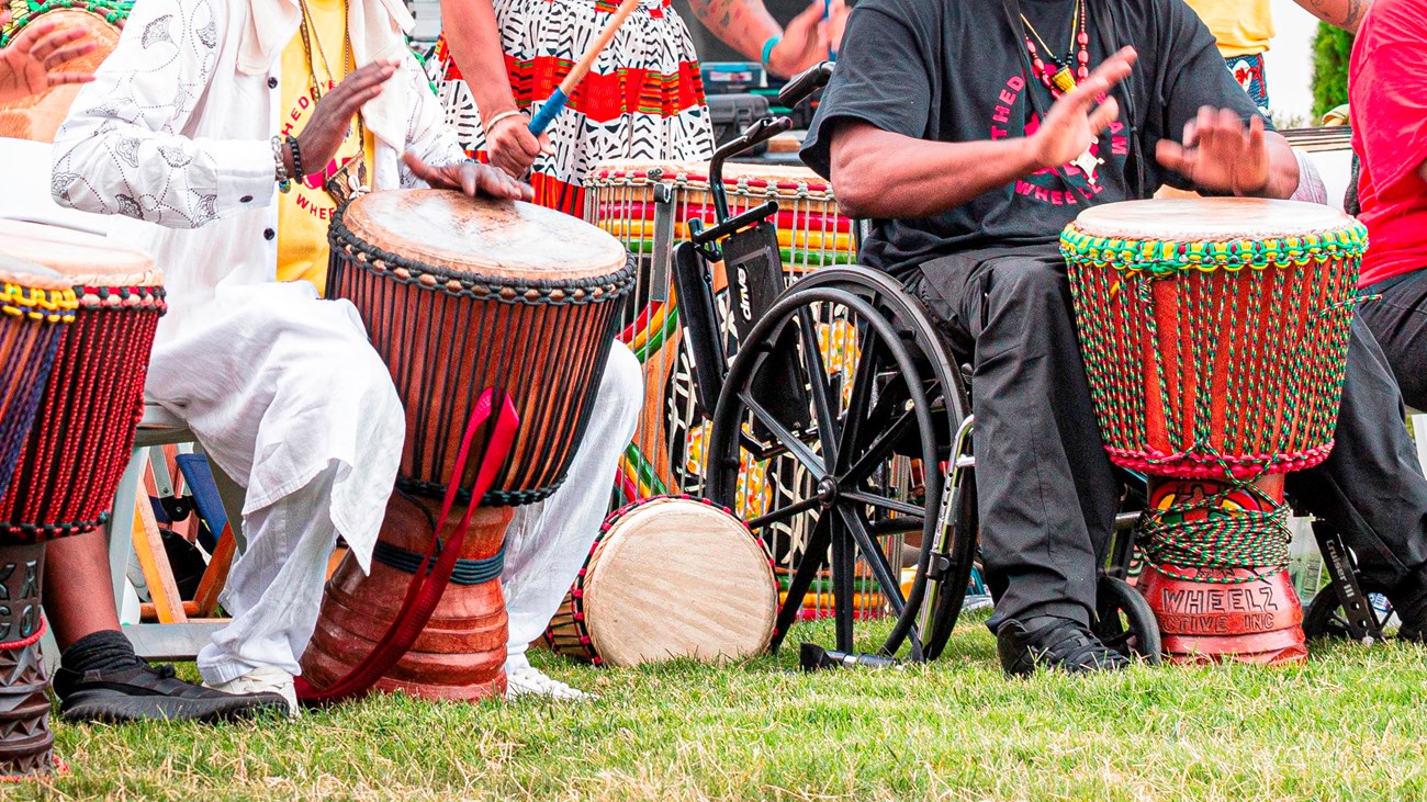 A line of colorfully dressed seated people in colorful clothes beat drums with their hands. .